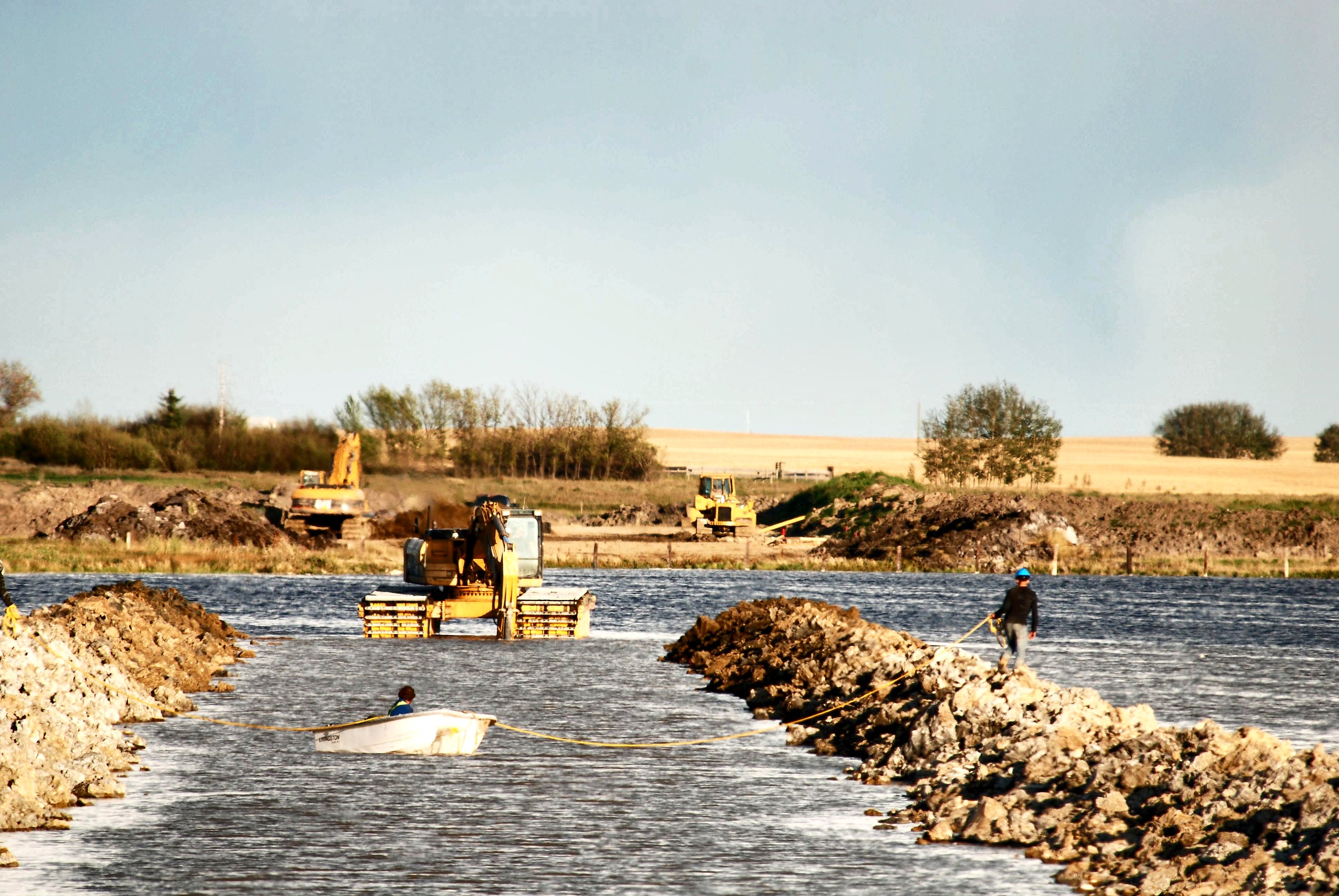 A crew, a boat and an amphibious excavator building a rock causeway across open water at Weed Lake, Alberta